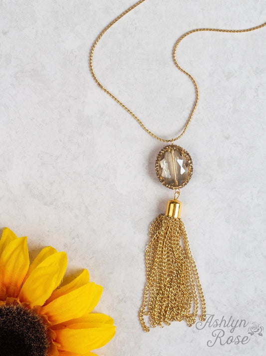 Gold tassel necklace with a large faceted gemstone pendant and layered gold chain fringe, styled on a light surface with a sunflower in the corner.