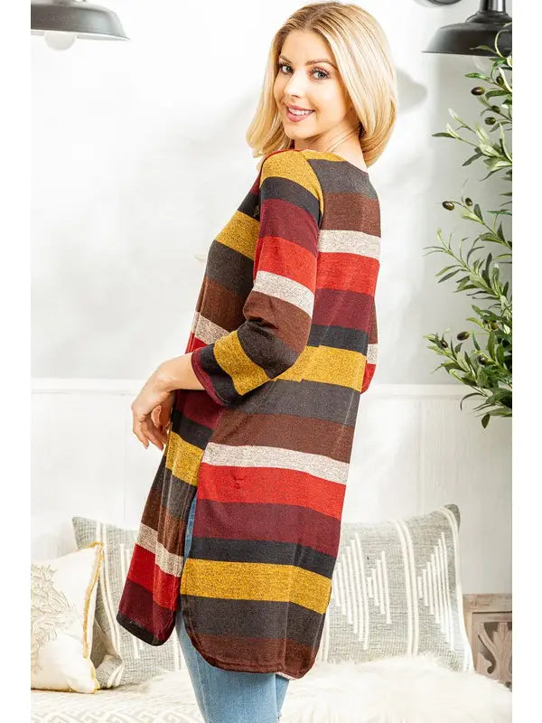 Woman smiling indoors while modeling a long, open-front cardigan with wide horizontal stripes in burgundy, mustard yellow, rust orange, cream, and charcoal. The cardigan has three-quarter sleeves and side slits, paired with light blue jeans.
