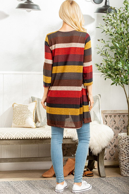 Back view of a woman wearing a long, open-front cardigan with wide horizontal stripes in burgundy, mustard yellow, rust orange, cream, and charcoal. The cardigan has three-quarter sleeves and side slits, styled with light blue skinny jeans and white sneakers.