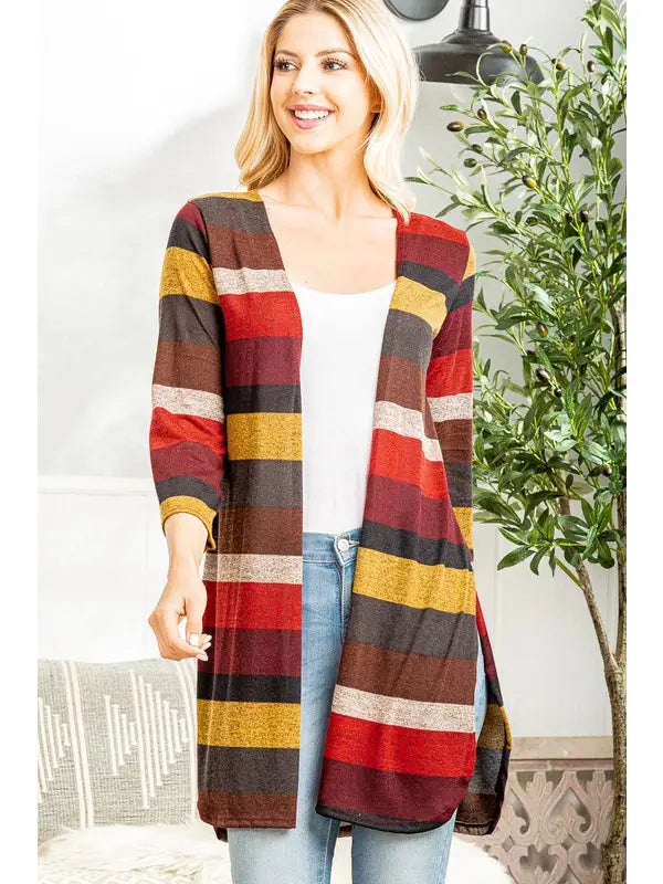 Woman indoors wearing a long, open-front cardigan with wide horizontal stripes in burgundy, mustard yellow, rust orange, cream, and charcoal. The cardigan features three-quarter sleeves and side slits, styled with a white tank top and light blue jeans.