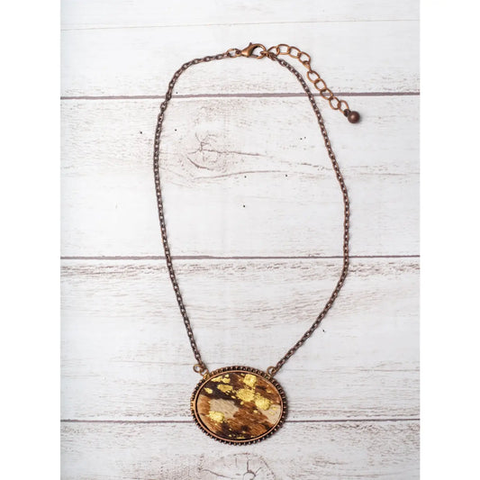 Oval cowhide pendant necklace with gold foil accents and vintage copper chain, displayed on white rustic wood background.