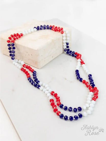 Red, white, and blue faceted bead double-strand necklace styled in a loose loop, displayed on a white marble surface with a cream-colored block and "Ashlyn Rose" logo in the corner.