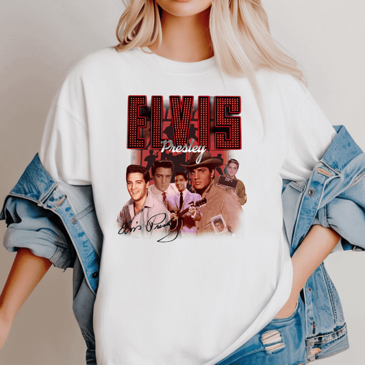 Woman wearing a white oversized T-shirt featuring a bold graphic collage of Elvis Presley in various iconic looks. The shirt has large red marquee-style text reading “ELVIS” with “Presley” in script underneath, surrounded by multiple vintage images of Elvis. His signature appears at the bottom of the design. She has layered the shirt with a light denim jacket, worn off the shoulders.