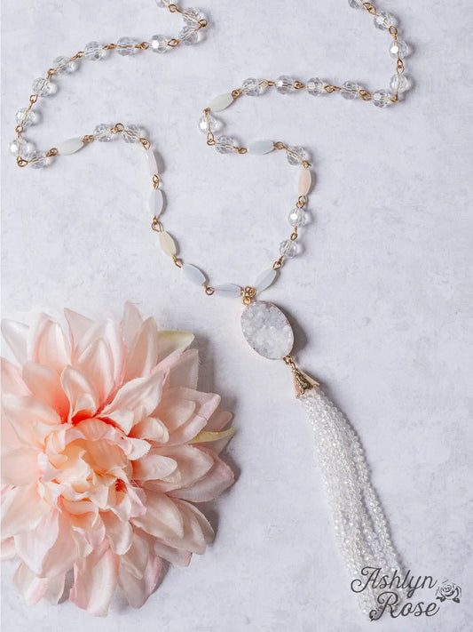 White druzy pendant necklace with crystal bead tassel, paired with a clear and opalescent beaded chain, styled next to a blush pink flower on a light background.