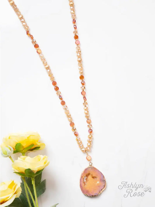 Beaded necklace in warm amber and peach tones with a shimmering druzy geode pendant, styled on a white background with yellow roses.