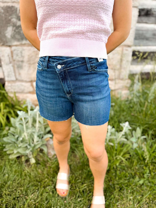 A woman wearing the CrissCharm Judy Blue Shorts, showcasing the high-waisted design with a criss-cross waistband and a diagonal front fly panel.