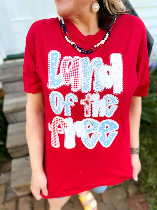 Woman wearing a bright red T-shirt that reads “Land of the Free” in large, playful patchwork-style letters featuring patriotic patterns like stars, gingham, and lace. She accessorizes with red, white, and blue beaded necklaces and pearl drop earrings, standing outdoors on a brick pathway near wooden steps.
