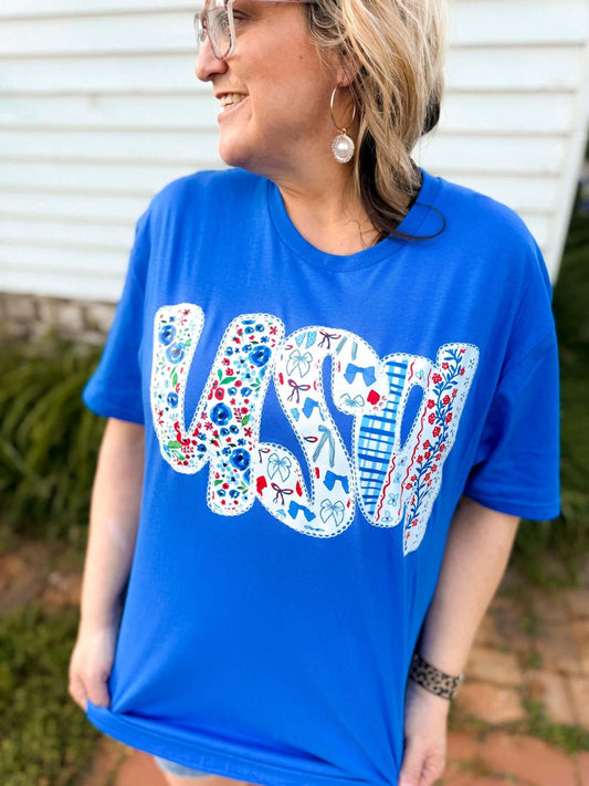 Woman wearing a royal blue T-shirt with the word “USA” in large, bold letters across the front. Each letter features a patchwork design of red, white, and blue patterns including florals, stars, gingham, and patriotic icons like sunglasses and fireworks. She accessorizes with pearl drop earrings, large hoop earrings, and glasses, smiling outdoors near a white house.