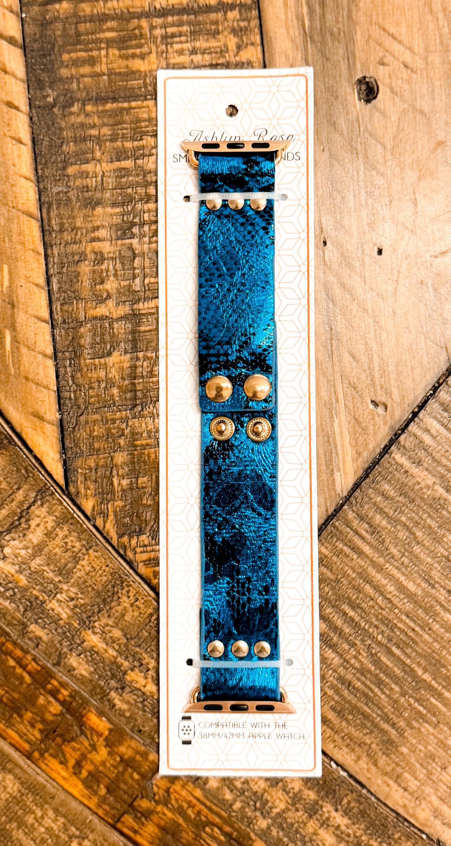 Metallic blue faux leather Apple Watch band with gold rivet snaps, displayed on product card with wooden background.