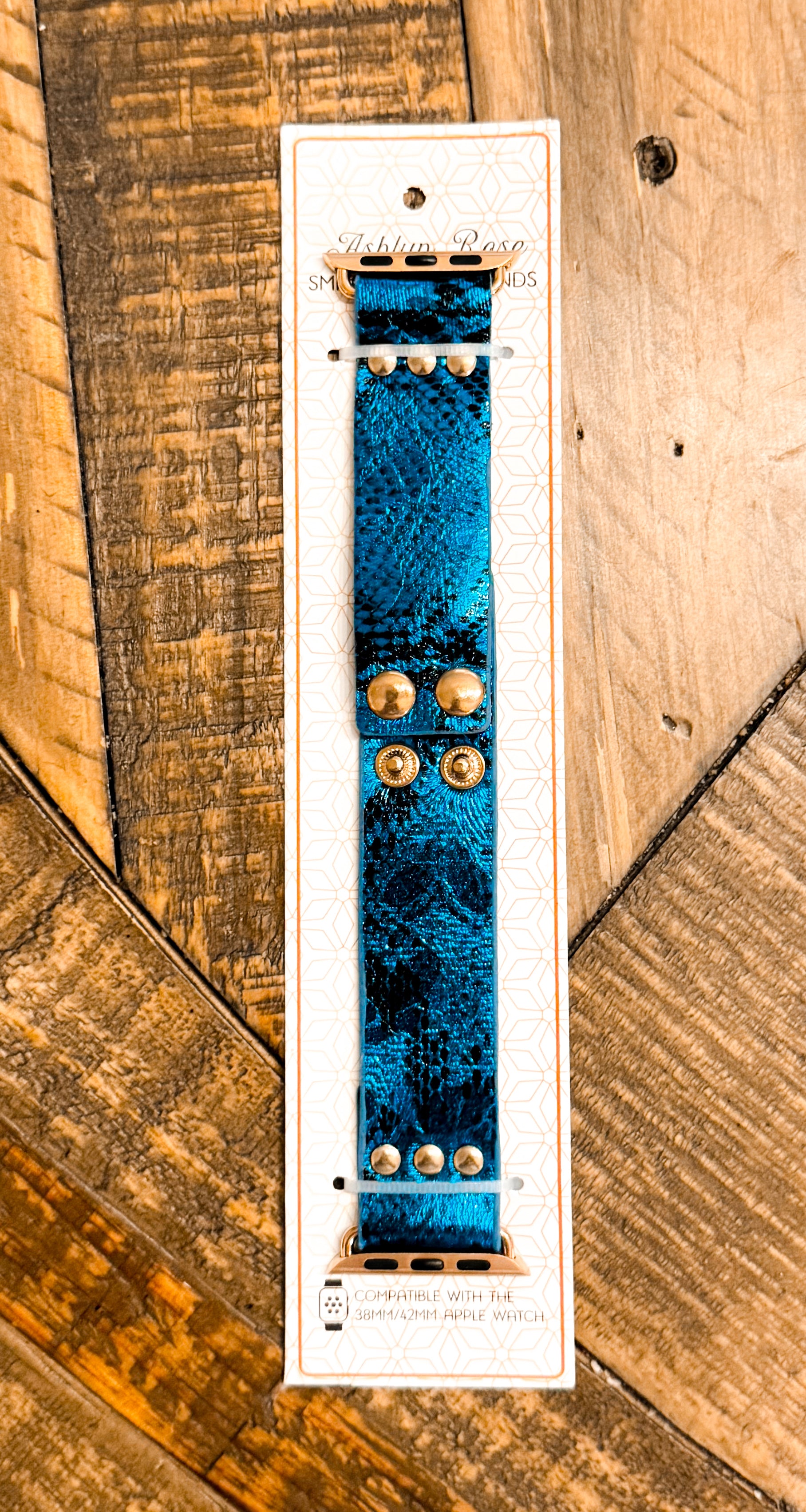 Metallic blue faux leather Apple Watch band with gold rivet snaps, displayed on product card with wooden background.