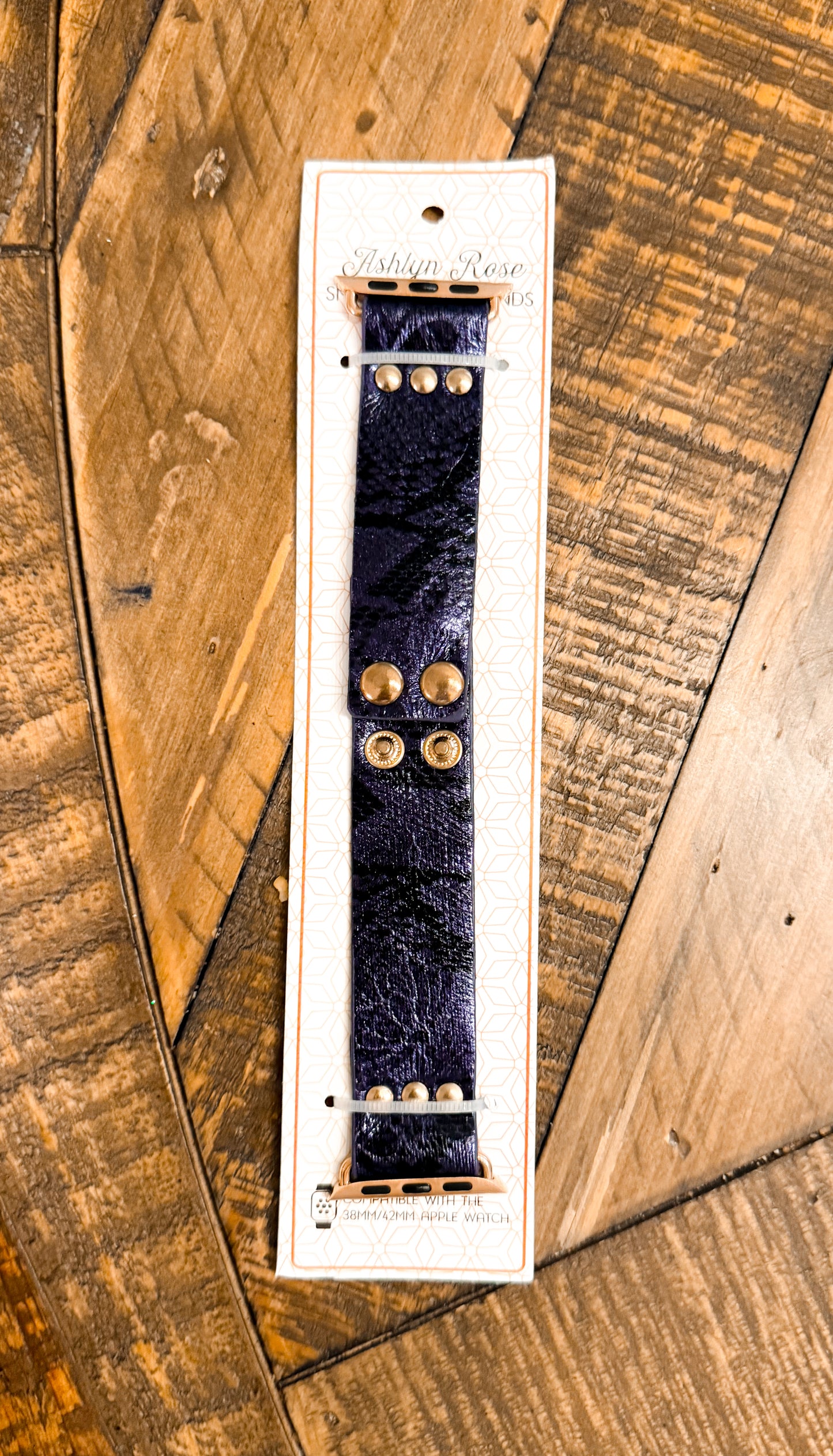 Shiny dark blue faux leather Apple Watch band with gold snap hardware, packaged on branded card atop rustic wood surface.