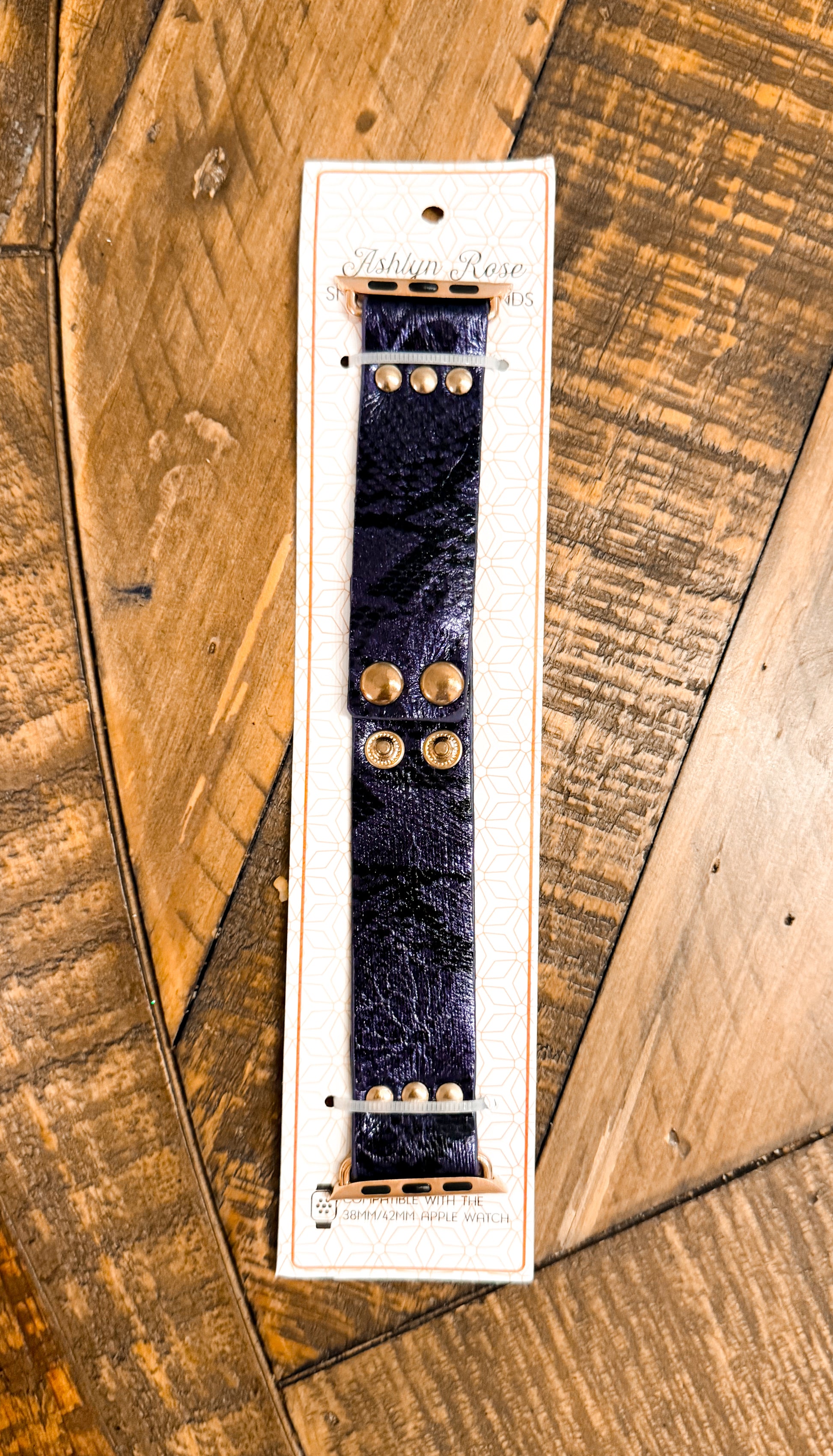 Shiny dark blue faux leather Apple Watch band with gold snap hardware, packaged on branded card atop rustic wood surface.