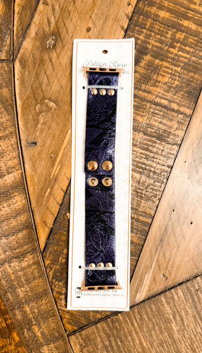 Shiny dark blue faux leather Apple Watch band with gold snap hardware, packaged on branded card atop rustic wood surface.
