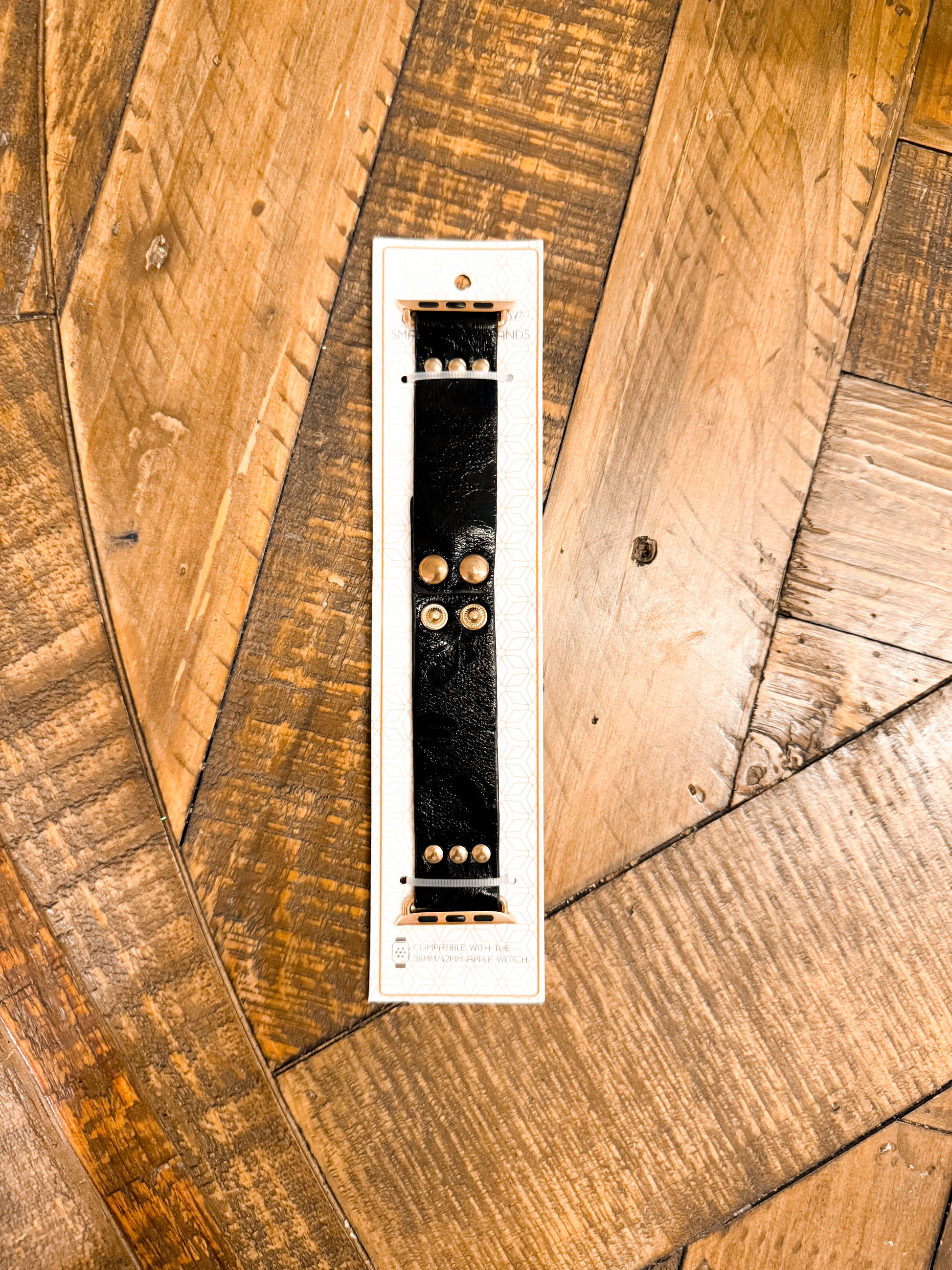 Glossy black faux leather Apple Watch band featuring gold snap buttons, laid on display card against a textured wood floor.