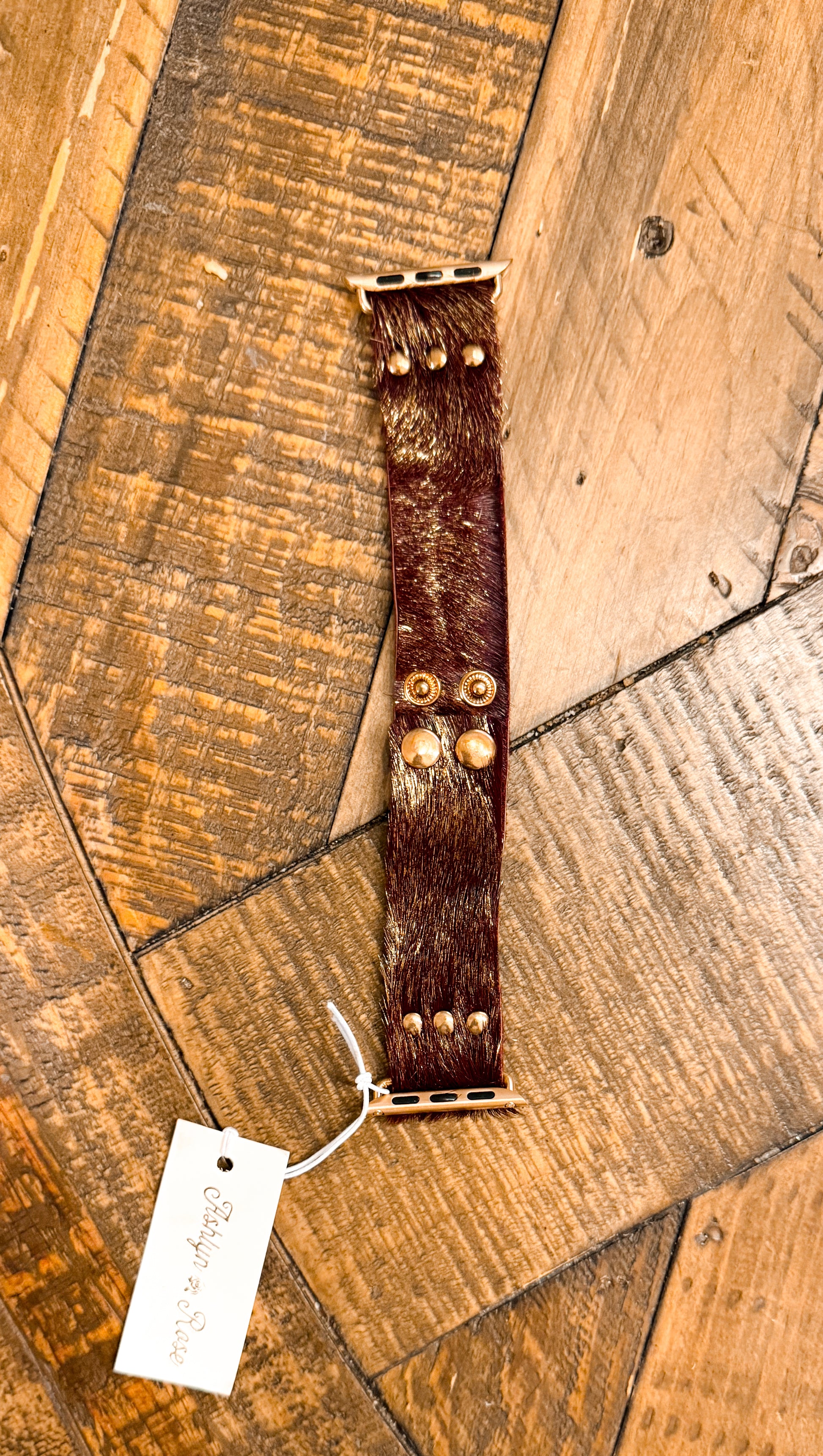 Brown and gold hair-on hide Apple Watch band with gold hardware, featuring visible texture and natural sheen on wooden background.