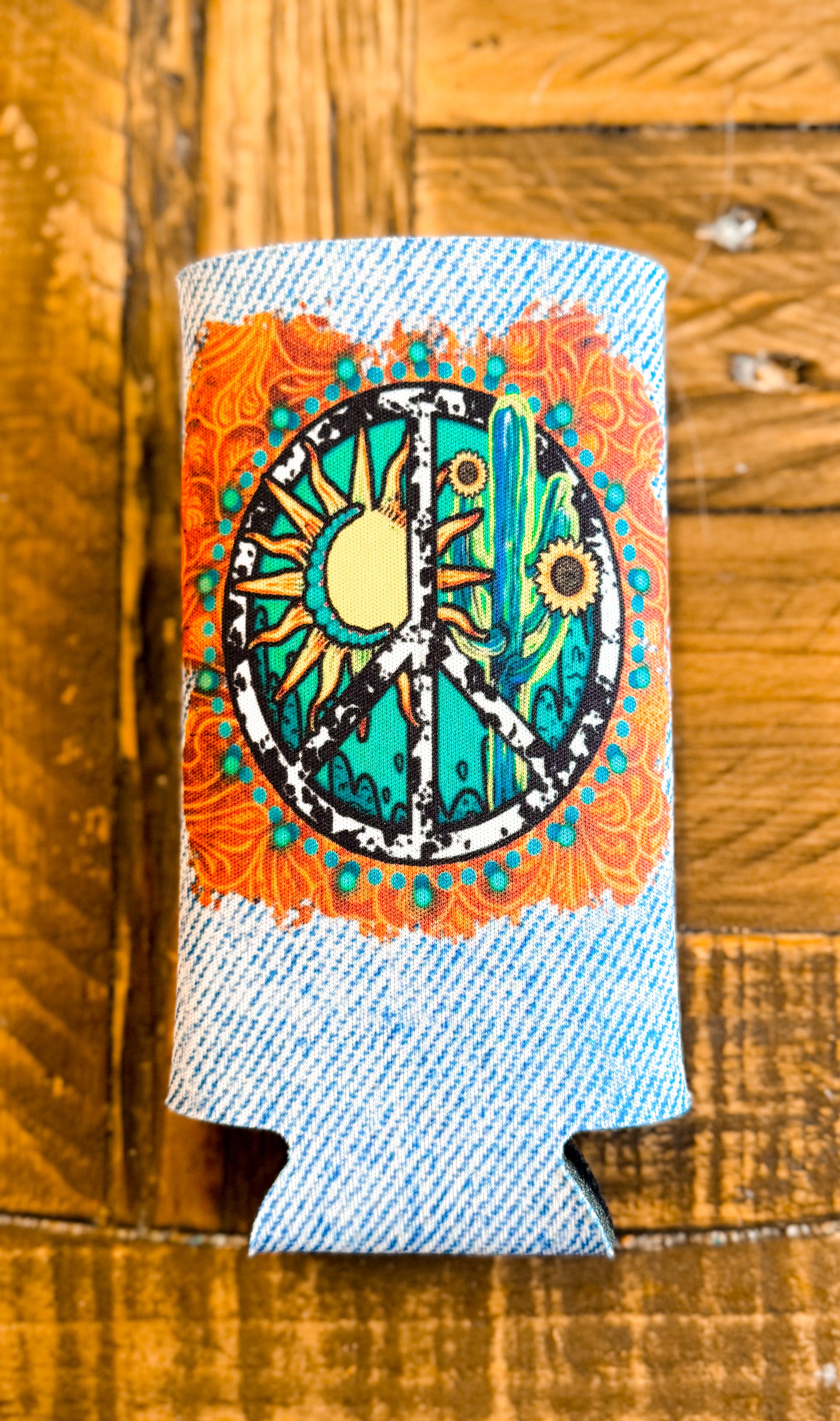 Slim can cooler with a denim-printed background and a vibrant peace sign design featuring a sun, cactus, and sunflowers surrounded by orange and turquoise mandala details.