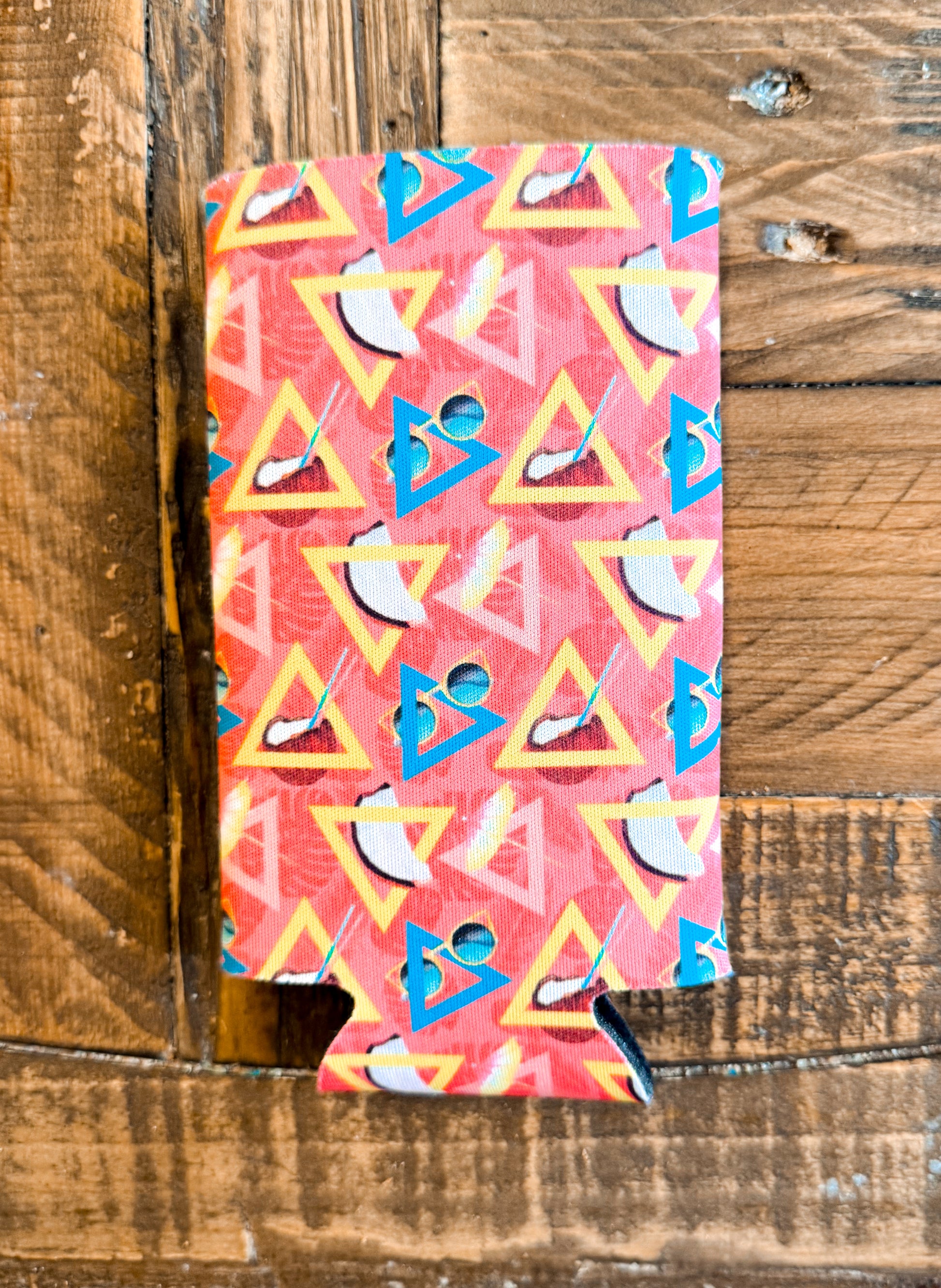 Slim can cooler with a bright red and coral geometric background featuring yellow triangles, cocktail glasses, lips, and beachy drink icons — totally 90s vacation vibes.
