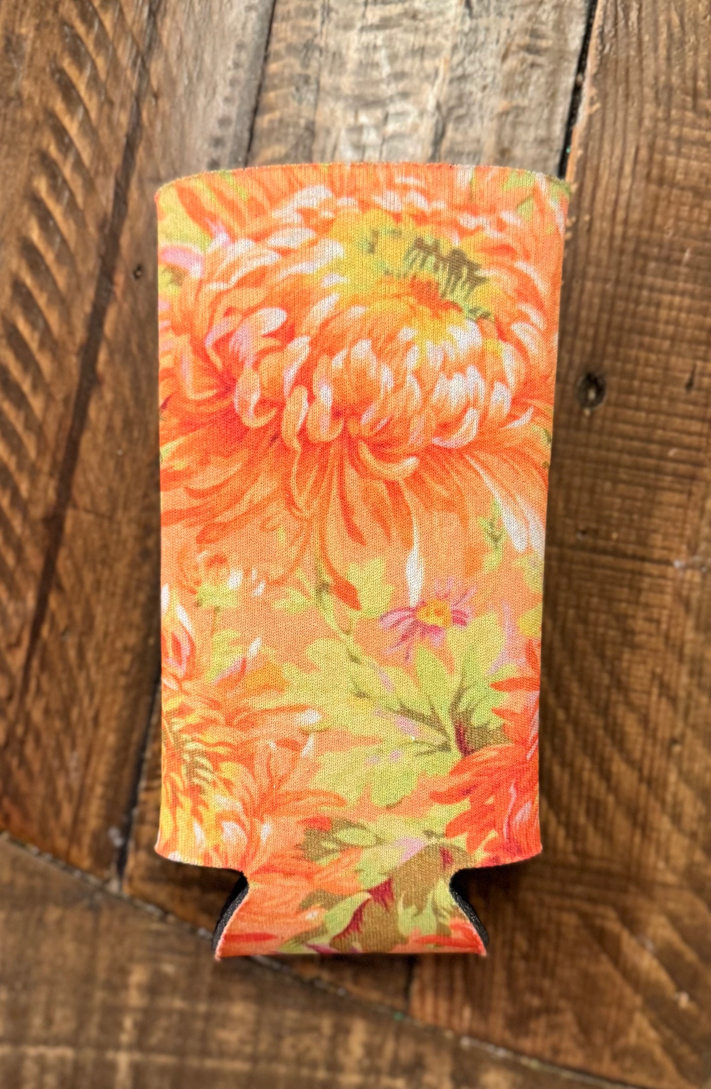 Slim can cooler with a vibrant floral pattern featuring shades of red, pink, purple, and orange flowers with dark centers, displayed on a rustic wooden floor background.