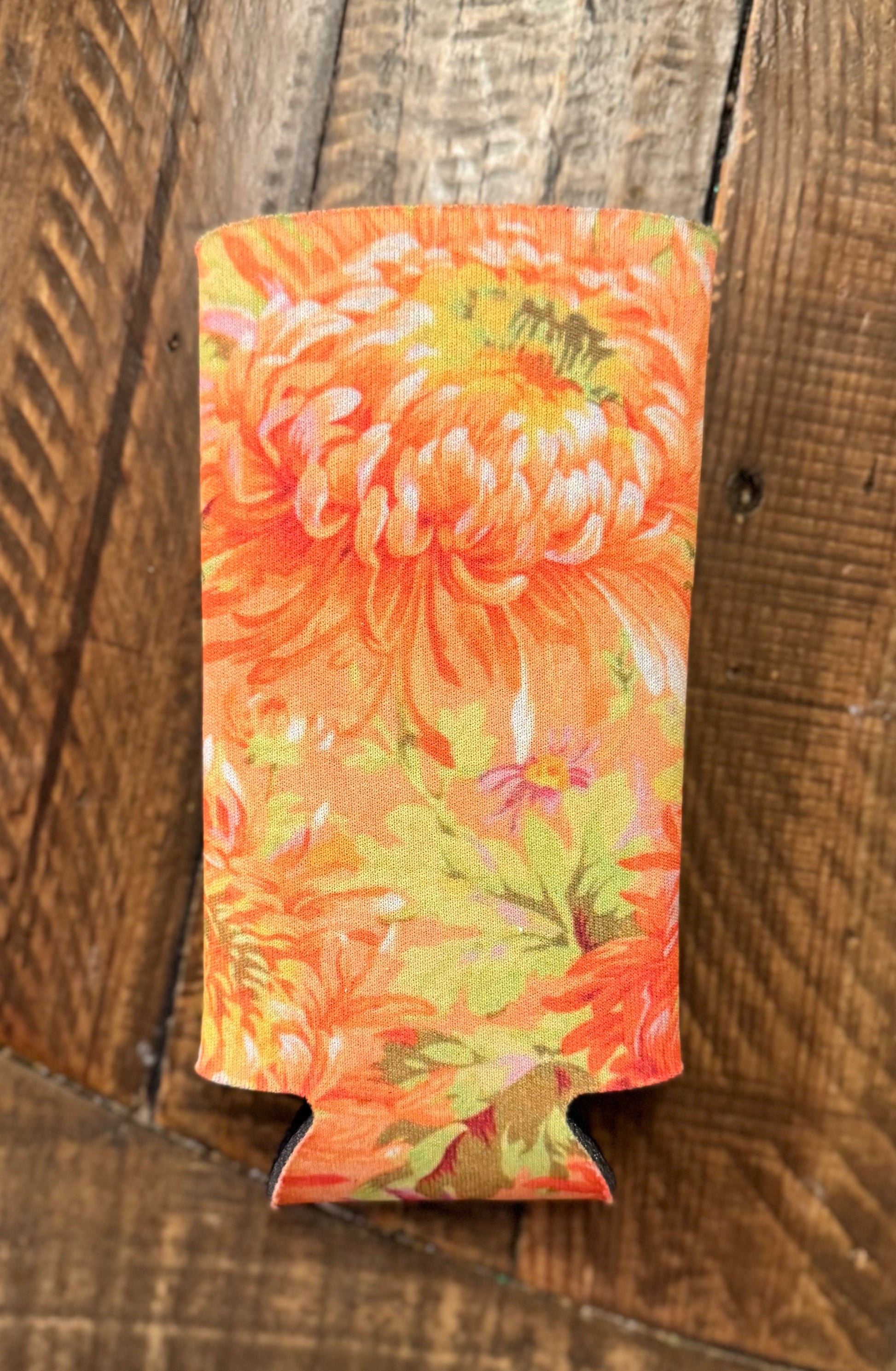 Slim can cooler with a vibrant floral pattern featuring shades of red, pink, purple, and orange flowers with dark centers, displayed on a rustic wooden floor background.