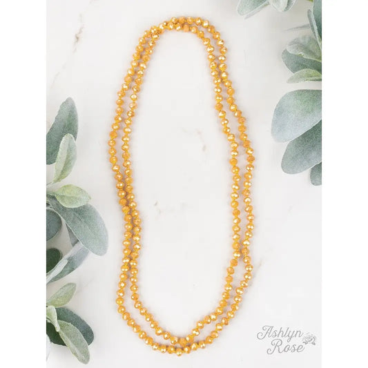Desert Marigold Necklace featuring mustard yellow faceted beads, styled as a long double-wrap strand, displayed on a white marble surface with soft green foliage accents.