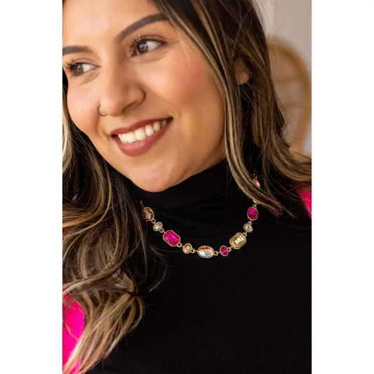Woman smiling while wearing the Glitz & Grace Necklace, featuring vibrant pink and iridescent gemstones set in gold, styled with a black turtleneck and hot pink accents.