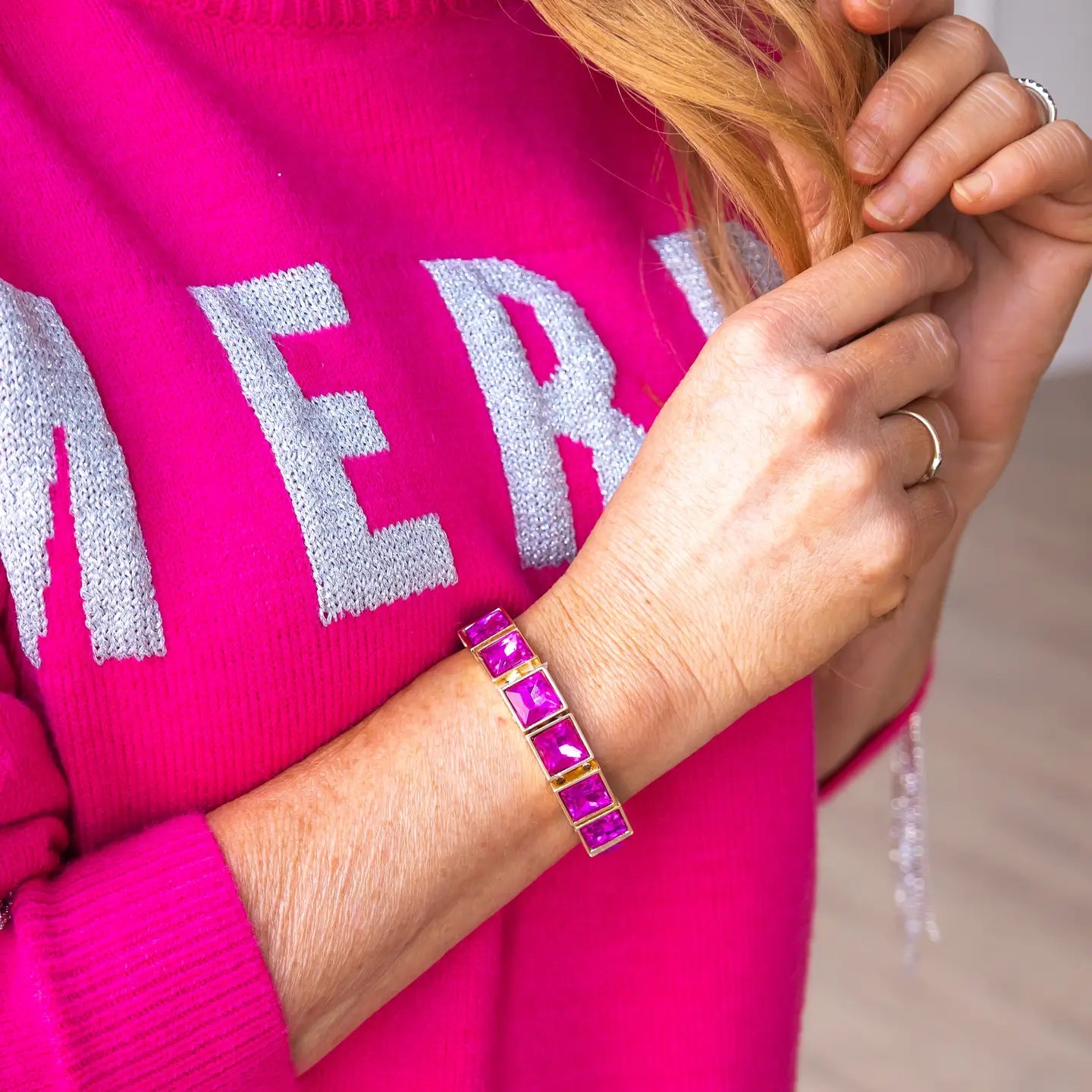 Close-up of a woman's wrist wearing the Power Pink Bracelet — a gold stretch bracelet with large square fuchsia stones — paired with a bright pink sweater featuring silver "MERRY" lettering.