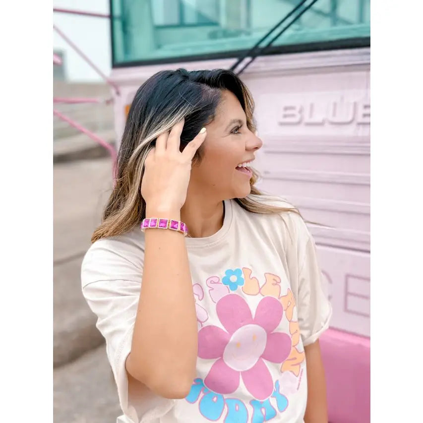 Smiling woman wearing the Power Pink Bracelet — a gold-toned stretch bracelet with bold square fuchsia stones — styled with a playful floral graphic tee, standing in front of a pastel pink bus.