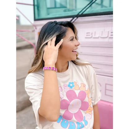 Smiling woman wearing the Power Pink Bracelet — a gold-toned stretch bracelet with bold square fuchsia stones — styled with a playful floral graphic tee, standing in front of a pastel pink bus.