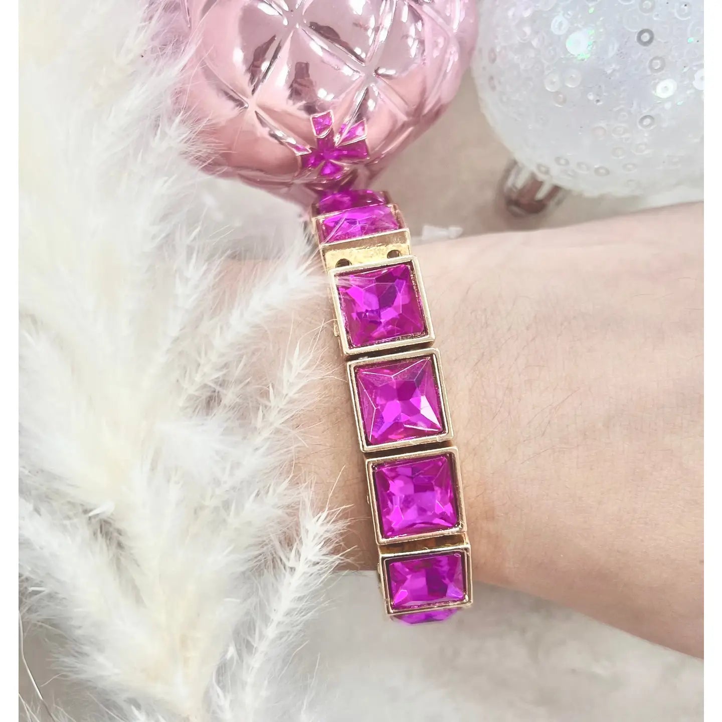 Close-up of the Power Pink Bracelet on wrist — featuring bold square fuchsia stones in gold settings — surrounded by soft white feathers and decorative pink and white ornaments.