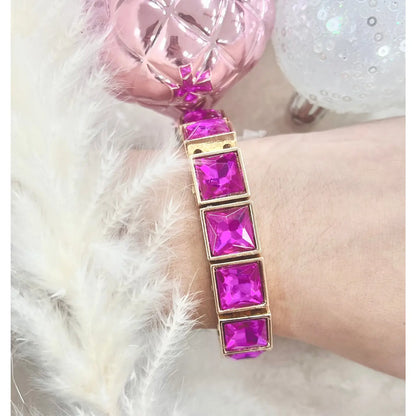 Close-up of the Power Pink Bracelet on wrist — featuring bold square fuchsia stones in gold settings — surrounded by soft white feathers and decorative pink and white ornaments.