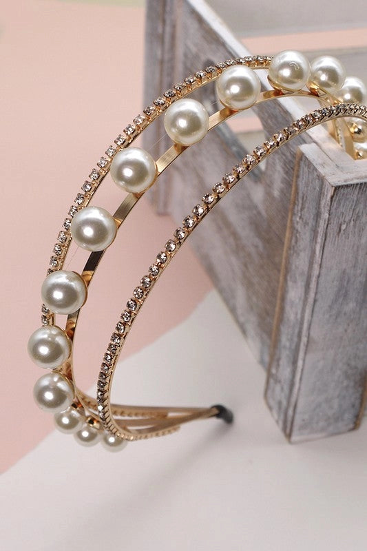 Gold double-band headband featuring large pearls and rows of rhinestones, styled on a pastel surface next to a wooden box. Elegant, feminine, and glam accessory.