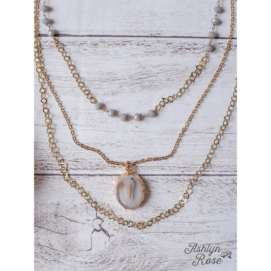 Layered gold necklace with milky gray beads and a white stone pendant, styled flat on a rustic white wood background with “Ashlyn Rose” logo.