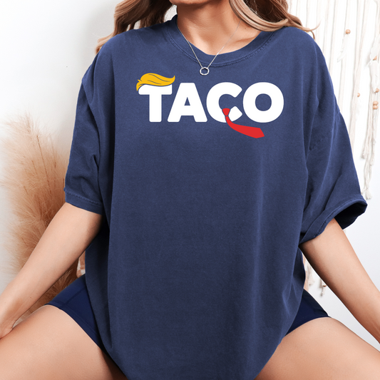 Woman wearing an oversized navy graphic t-shirt with bold white "TACO" text, featuring a yellow hair graphic on the "T" and red tie on the "C," sitting cross-legged in a boho-inspired setting.