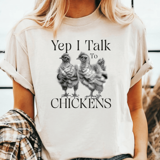 Woman wearing a natural-colored T-shirt with rolled sleeves, featuring a black and white sketch-style graphic of two fluffy chickens. Above and below the chickens, the text reads “Yep I Talk To CHICKENS” in a mix of serif fonts. She is styled casually with loose waves and a flannel shirt draped over one arm.