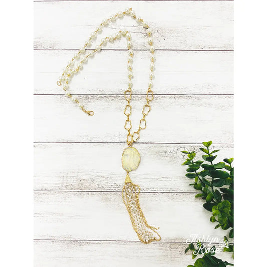 Long pearl and gold chain necklace with a shimmering oval pendant and layered tassel fringe, styled on a white wood background with greenery.