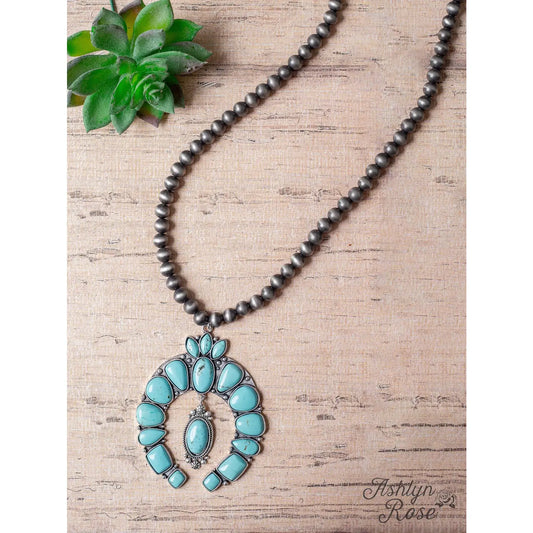 High Desert Bloom Necklace — squash blossom-style necklace with large turquoise stones set in antique silver and strung on oxidized silver beads — displayed on a rustic wood background with a green succulent accent.